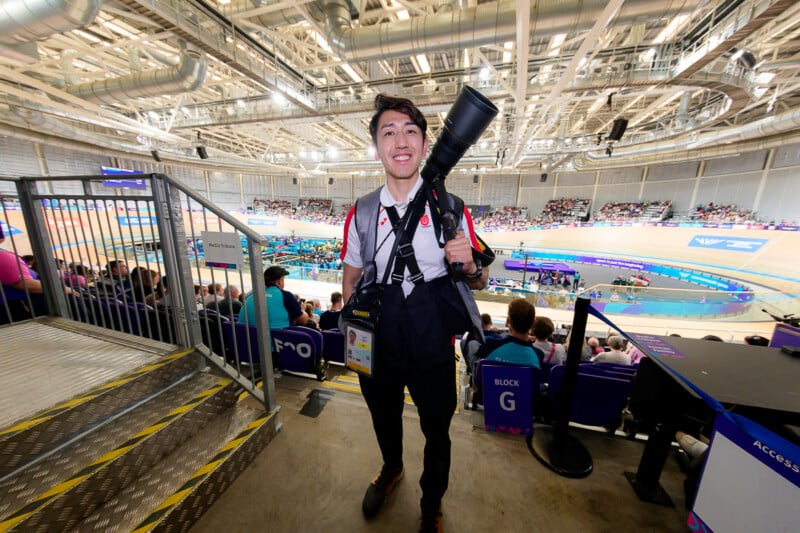 A smiling photographer stands in a stadium media section, holding a large telephoto lens over his shoulder while wearing a camera harness.