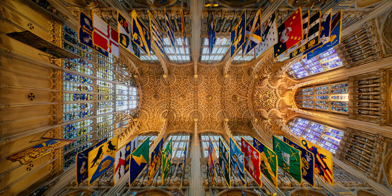 A low-angle view of an ornate, vaulted stone ceiling in a cathedral, featuring intricate fan vaulting and rows of colorful heraldic banners hanging from the walls.