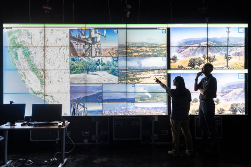 Two people stand before a large wall of monitors displaying maps and live camera feeds of landscapes, with one person pointing at a screen showing smoke.