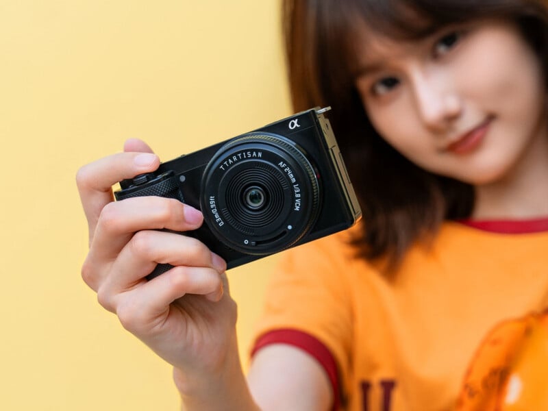 A person with shoulder-length brown hair holds a black digital camera with a TTArtisan AF 24mm f/1.8 VCM lens toward the viewer against a yellow background.