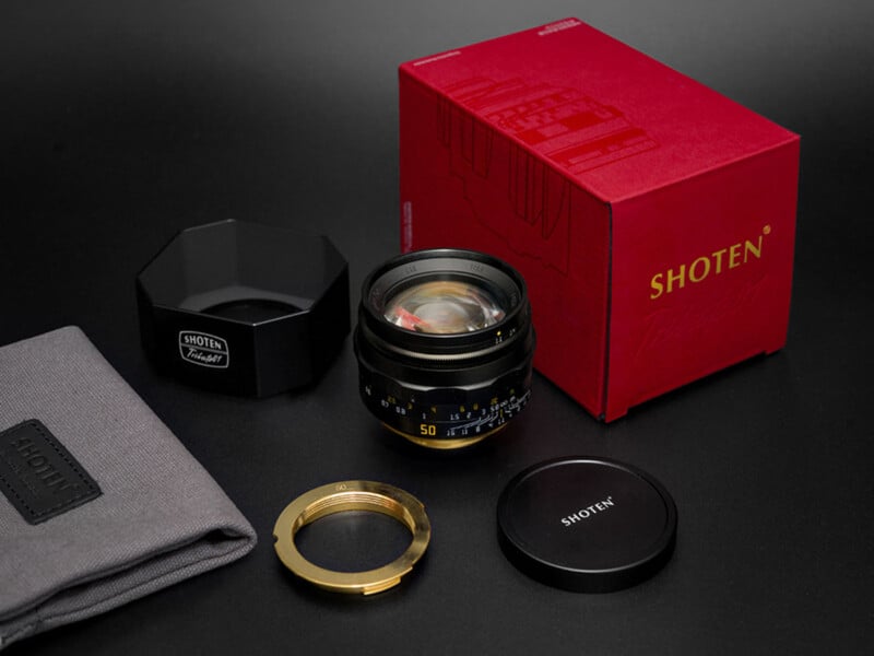 A Shoten Tribute 01 camera lens, its red box, a lens hood, a lens cap, a brass ring, and a grey storage pouch arranged on a black surface.