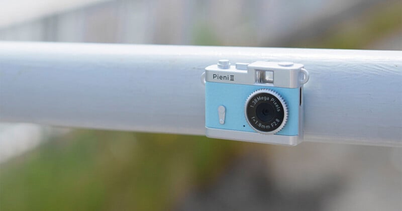 A small, light blue Pieni III toy-style digital camera is resting on a white horizontal railing.