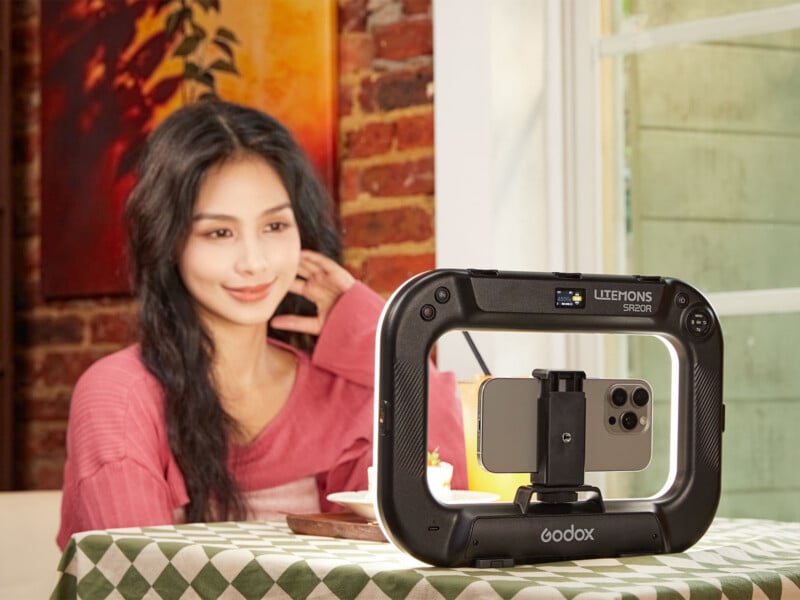 A woman in a pink shirt sits at a table behind a Godox Litemons SR20R ring light that holds a smartphone.