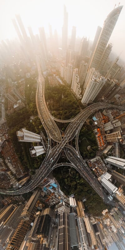 An aerial, wide-angle view of a multi-level highway interchange surrounded by dense city skyscrapers and patches of trees.