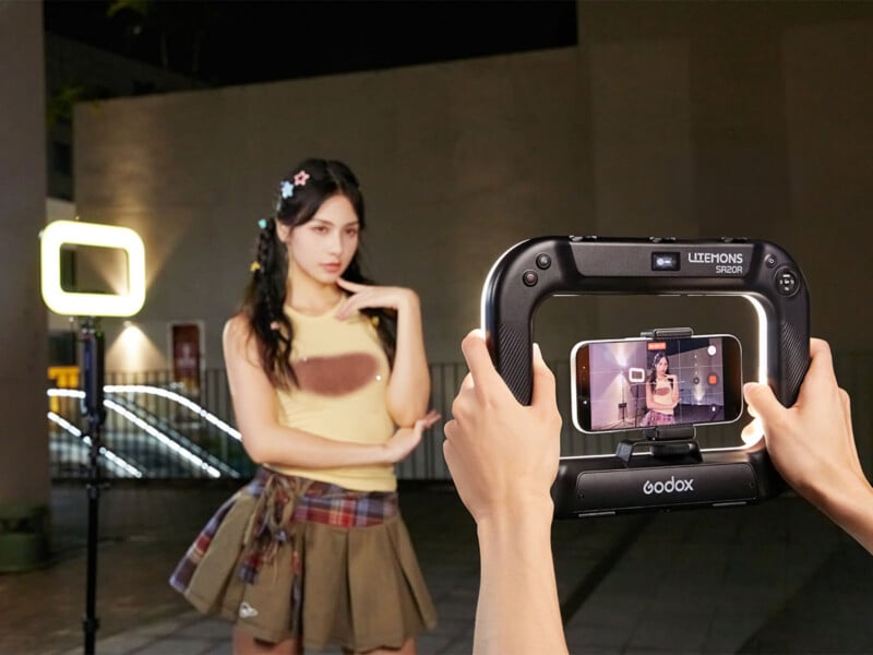 A person holds a Godox Litemons SR20R ring light frame with a smartphone mounted in the center, filming a young woman posing in a yellow tank top and pleated skirt.