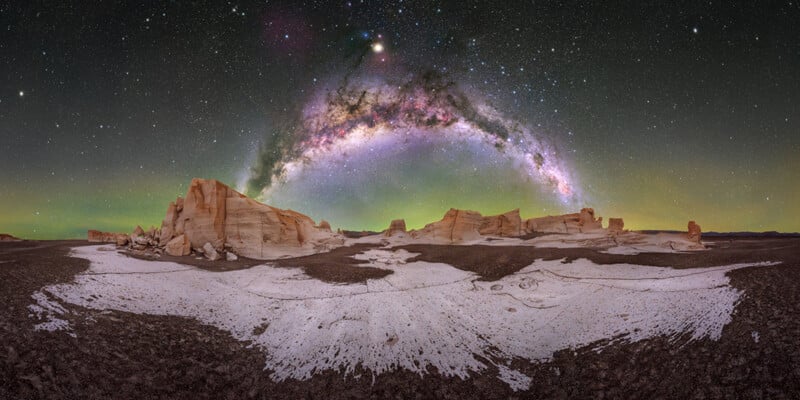 A panoramic view of a desert landscape with white rock formations under a vibrant, arched Milky Way galaxy and a starry night sky.