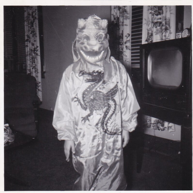 A child stands in a living room wearing a dragon mask and a costume featuring a large dragon illustration, with a vintage television set visible in the background.