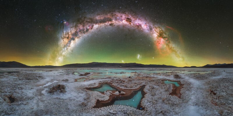 A panoramic view of the Milky Way arching over a salt flat with turquoise pools and distant mountains under a starry night sky.