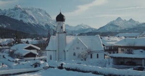 A snow-covered village with a white church featuring a dark onion dome sits in front of a backdrop of jagged, snow-capped mountains.