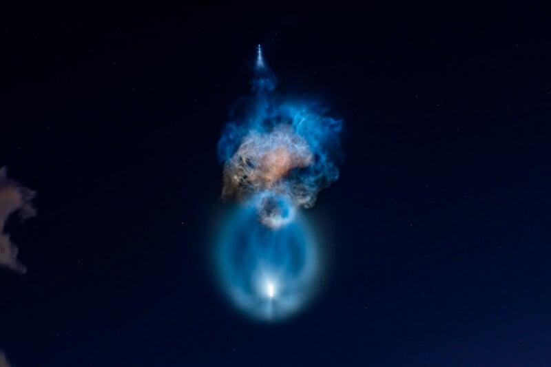 A bright rocket plume expands in the night sky, creating a glowing blue ring and a large, diffuse cloud of exhaust.