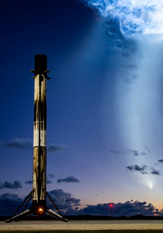 Falcon 9 rocket stands beside its launch tower while a diffuse exhaust cloud glows in the sky behind the pad.