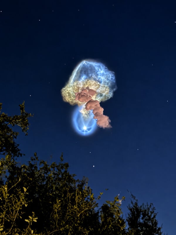 A bright, glowing blue and pink plume of exhaust from a rocket launch rises into a dark night sky above silhouetted trees.
