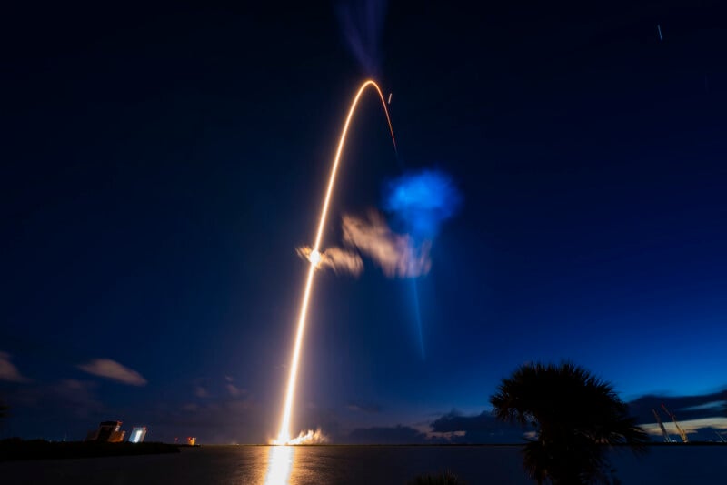 A rocket launches into the night sky, leaving a long, bright orange trail that curves toward the right, next to a blue cloud.