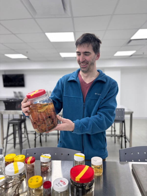 A man in a blue fleece jacket holds a large glass jar containing preserved specimens in a laboratory setting with several other sample jars on the table in front of him.