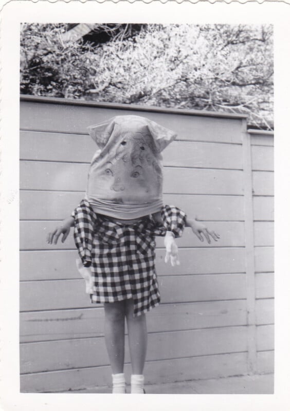 A person wearing a paper bag mask with a hand-drawn face and a plaid shirt stands in front of a wooden fence.
