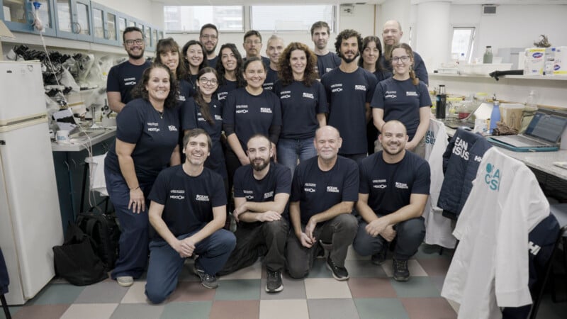A group of 19 people wearing matching navy blue Ocean Census t-shirts pose for a group photo in a laboratory setting.