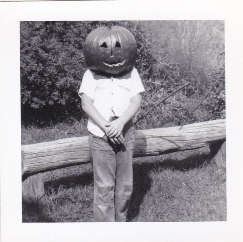 A child stands in front of a log, wearing a carved pumpkin as a mask over their head.