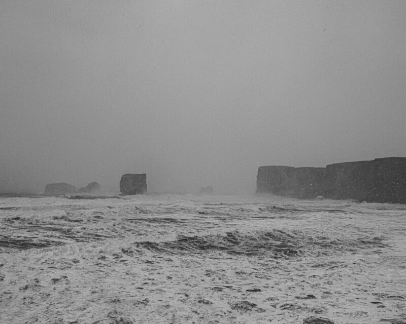 A black and white view of a turbulent, choppy ocean with large rock formations rising from the water under a snowy sky.