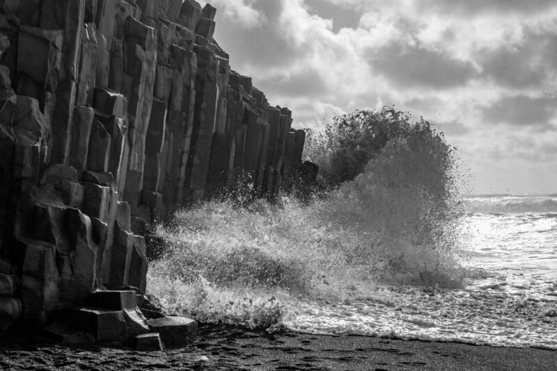 A large wave crashes against a dark, vertical basalt cliff on a black sand beach under a cloudy sky.