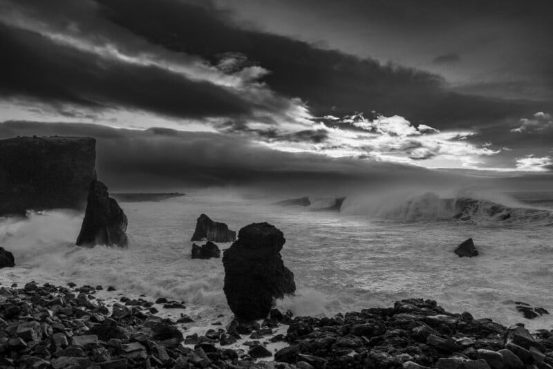 A black and white landscape of a stormy, turbulent ocean crashing against jagged dark rock formations and a rocky shoreline.