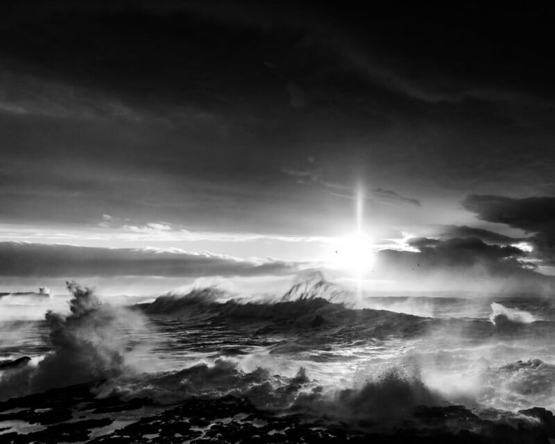 A black and white view of a turbulent ocean with large, crashing waves under a bright, sunlit sky with dramatic clouds.