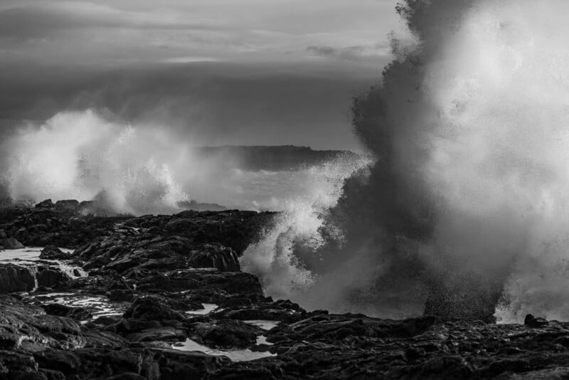 Large waves crash against a dark, jagged rocky coastline under a cloudy sky.
