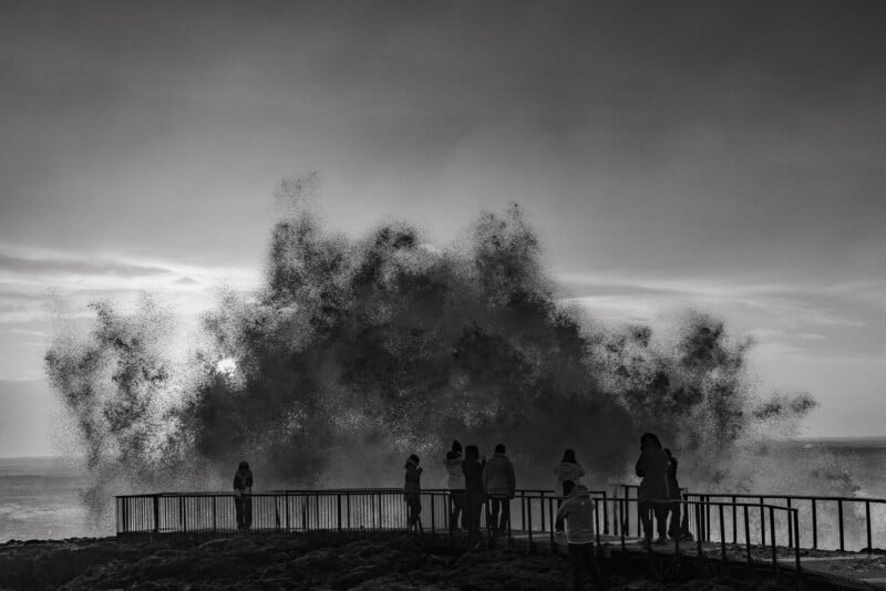 A group of people stands on a railed observation deck watching a large wave crash against the shore at sunset.