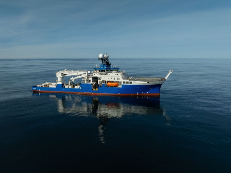 A blue and white research vessel, the Falkor (too), floats on calm, dark blue water under a clear sky.