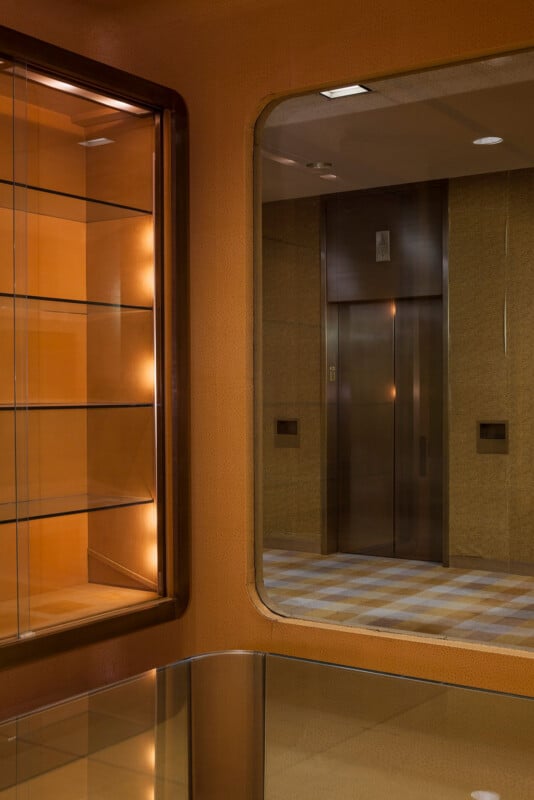 A glass display case sits next to a large mirror reflecting an elevator lobby with a metallic elevator door and a checkered floor.