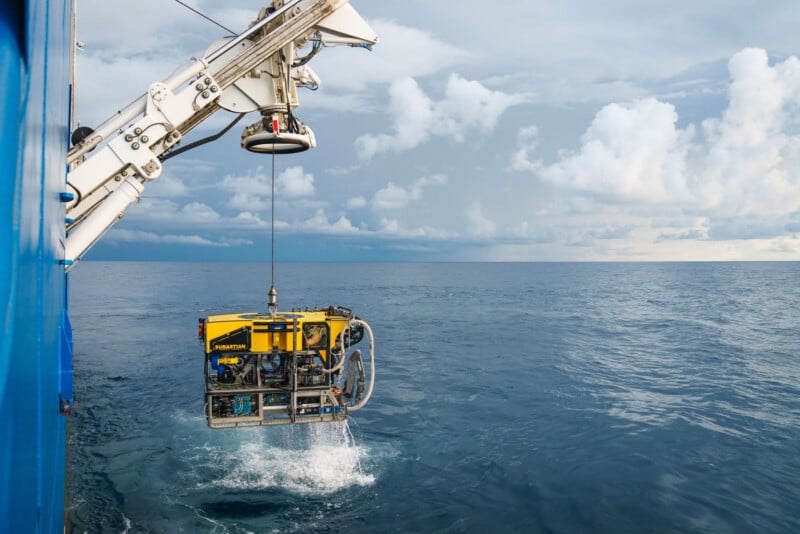 A yellow remotely operated vehicle named Subastian is being lowered into the ocean by a large white crane on a ship.