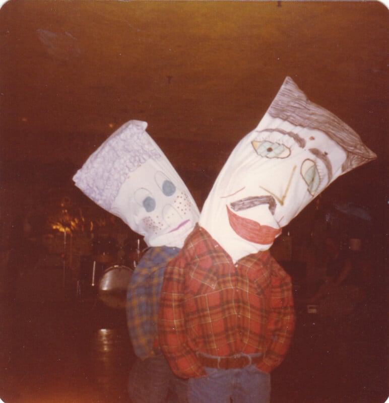 Two people stand side-by-side wearing large, hand-drawn paper bag masks over their heads and plaid flannel shirts.