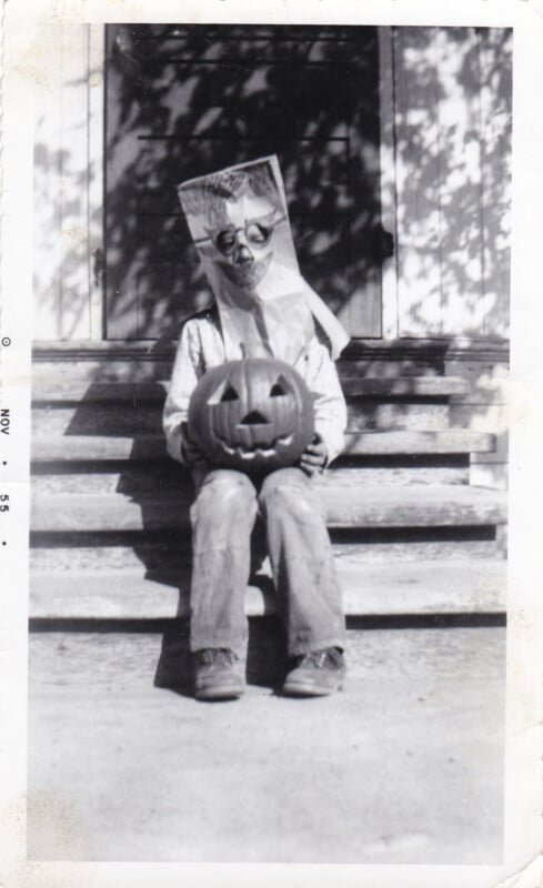 A child sits on porch steps wearing a paper bag mask with a hand-drawn face, holding a carved jack-o'-lantern.