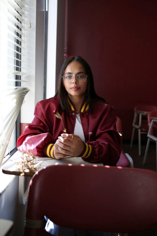 A young person with glasses and a maroon varsity jacket sits at a desk by a window, holding a dried flower.