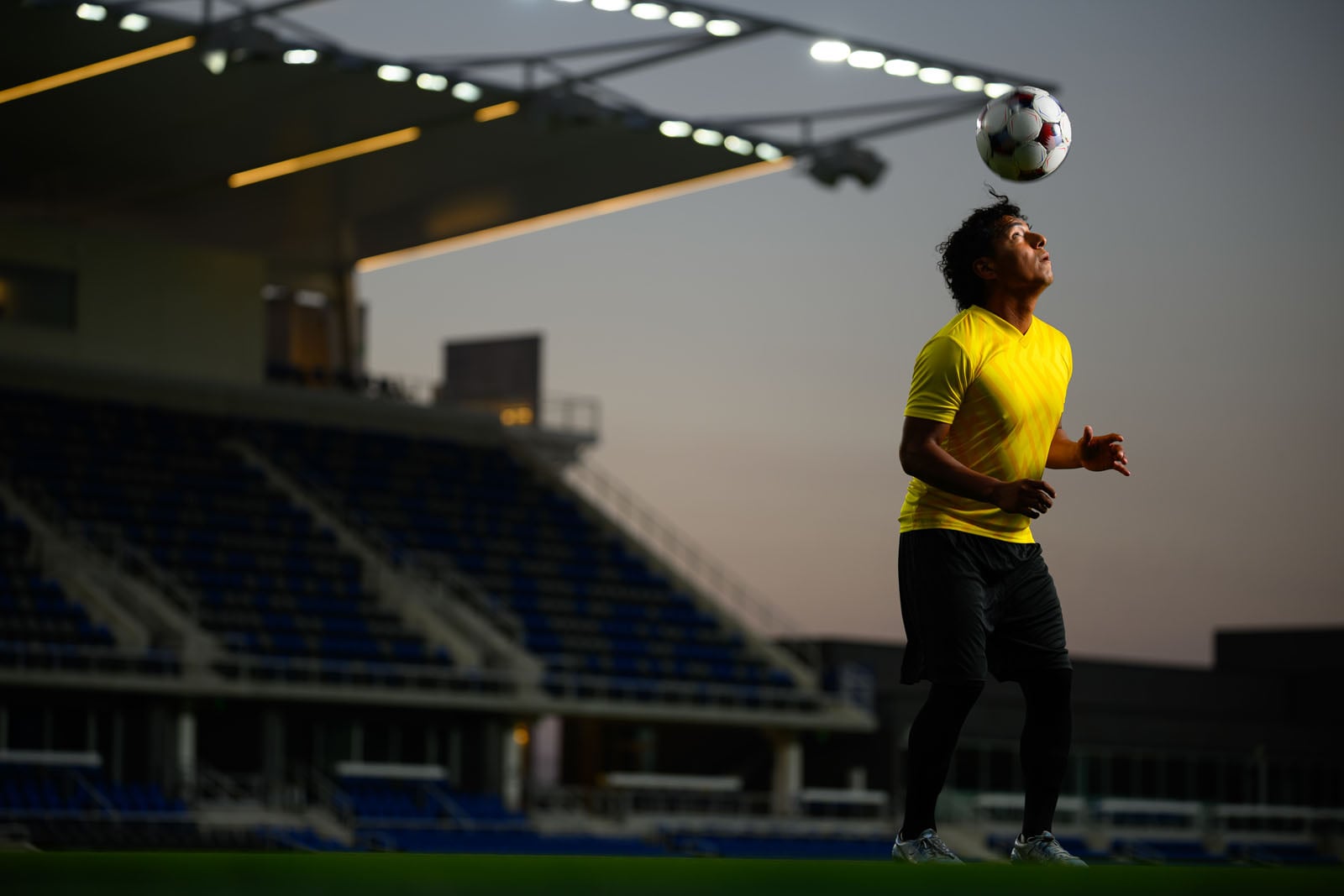 A soccer player in a yellow jersey and black shorts balances a soccer ball on his head at a stadium during twilight.