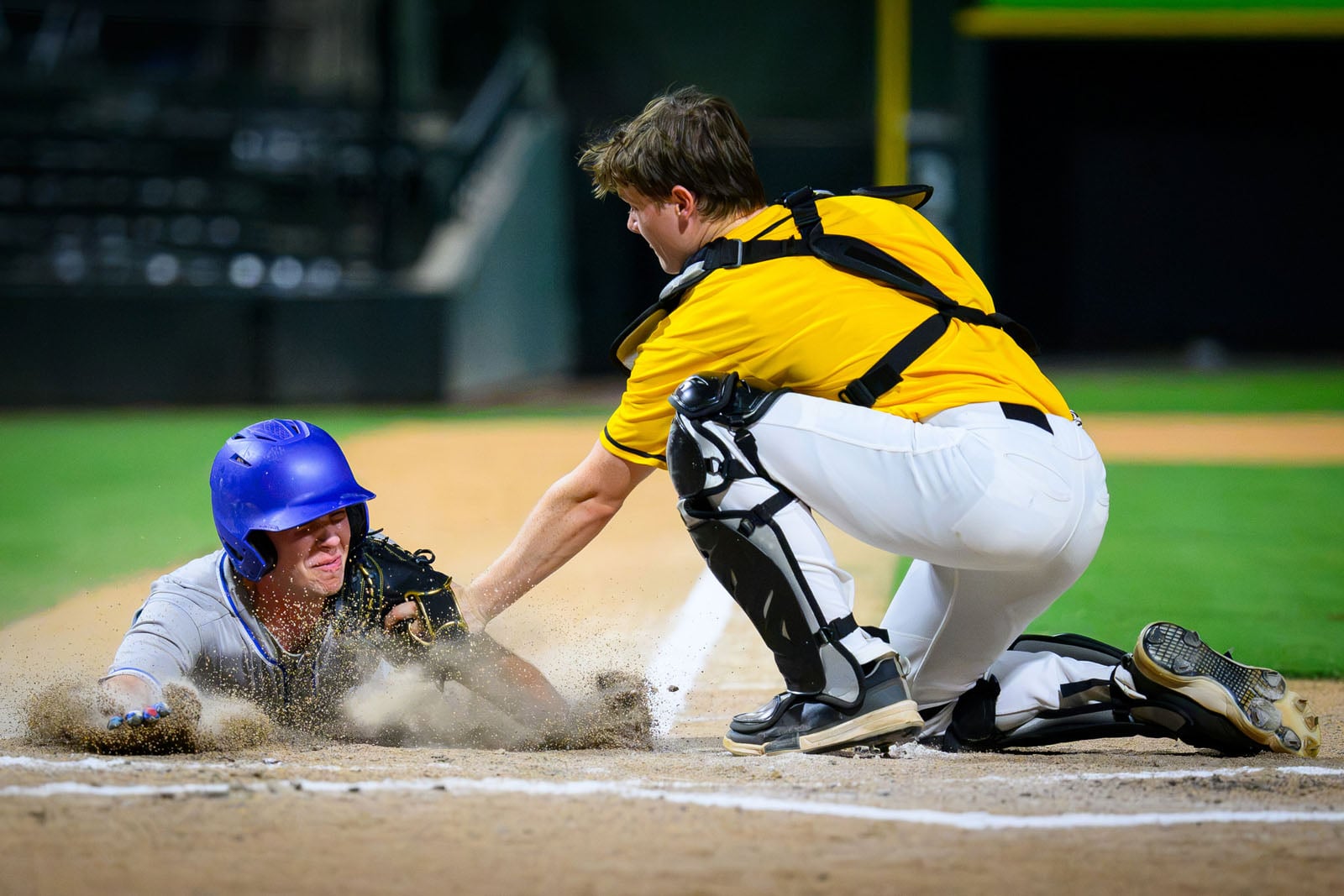 A baseball catcher in a yellow jersey tags a sliding runner wearing a blue helmet at home plate.