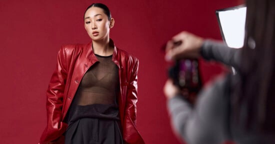 A model with slicked-back hair wears a red leather jacket over a sheer black top, posing against a red background while being photographed.