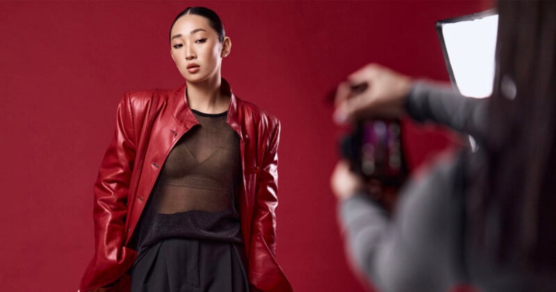 A model with slicked-back hair wears a red leather jacket over a sheer black top, posing against a red background while being photographed.