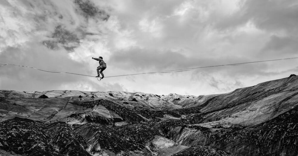 A person balances on a slackline stretched across a vast, rugged, and rocky mountain landscape under a cloudy sky.