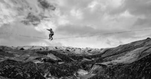 A person balances on a slackline stretched across a vast, rugged, and rocky mountain landscape under a cloudy sky.