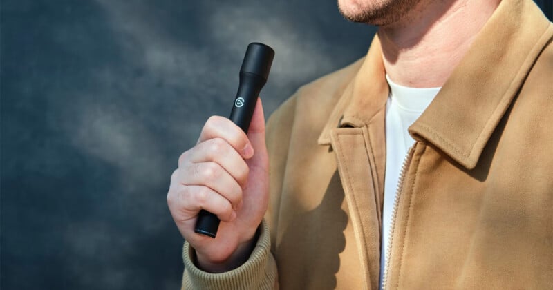 A person wearing a tan jacket holds a black, cylindrical handheld device with a small logo in their right hand.