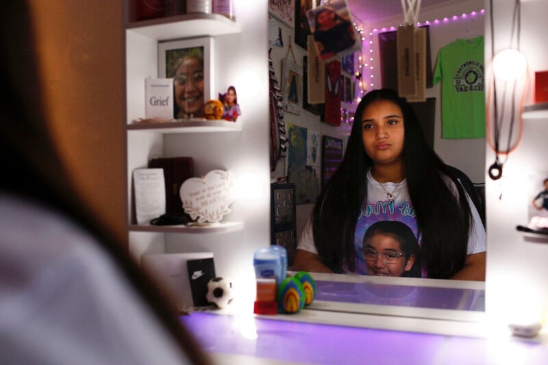 A young woman with long dark hair sits at a vanity mirror, wearing a shirt featuring a portrait of a girl in glasses.