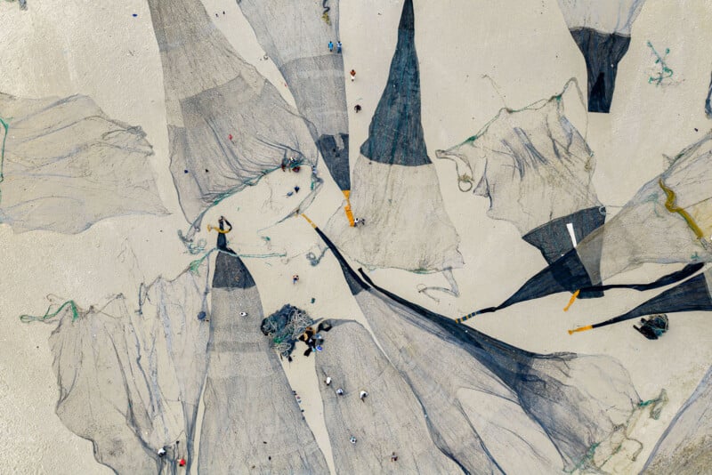 An aerial view shows several large, dark fishing nets spread out across a light-colored sandy beach, with small figures of people working among them.