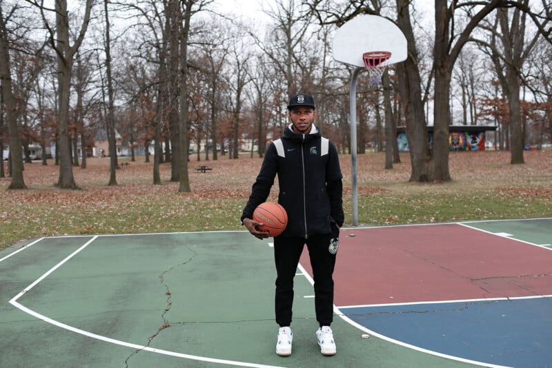 A man wearing a black hoodie, black pants, and a baseball cap stands on an outdoor basketball court holding a basketball.