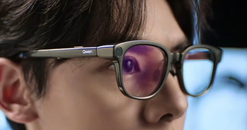 A close-up side view of a person wearing dark-rimmed glasses with the word "Qwen" printed on the temple.