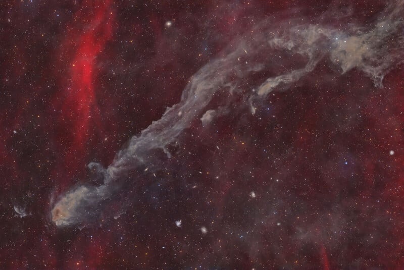A wispy, translucent nebula stretches diagonally across a deep red, star-filled field, with several small spiral galaxies visible in the background.