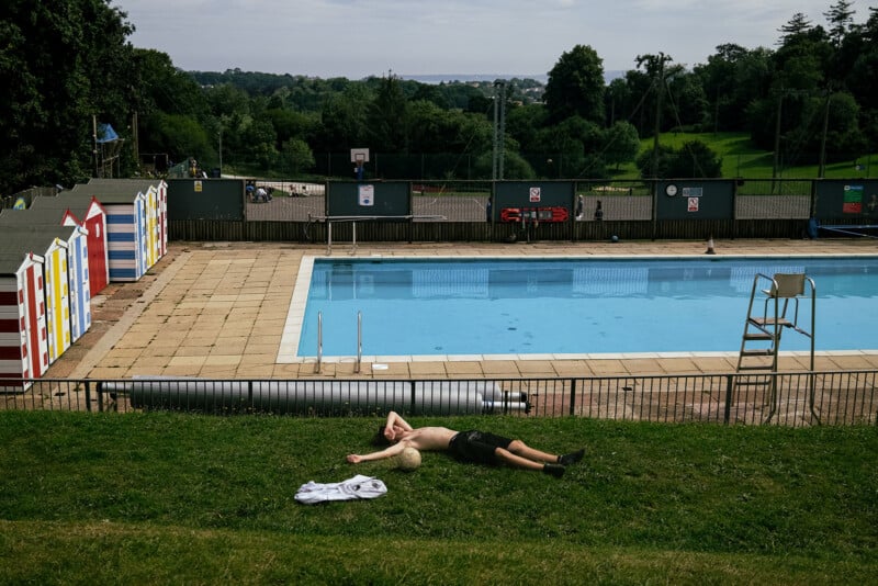 A shirtless man lies on the grass next to a soccer ball and a white shirt, with a swimming pool and colorful beach huts in the background.