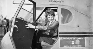 Amelia Earhart sits in the cockpit of a Hammond Y aircraft, holding the open door, with Department of Commerce markings visible on the side.