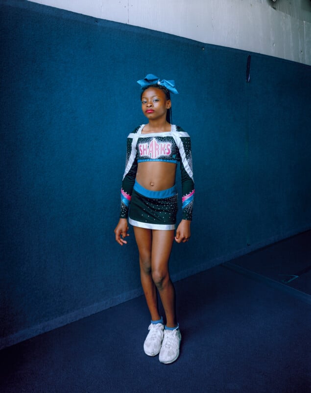 A young girl in a black and teal cheerleading uniform with "SHARKS" on the top stands against a dark blue wall.
