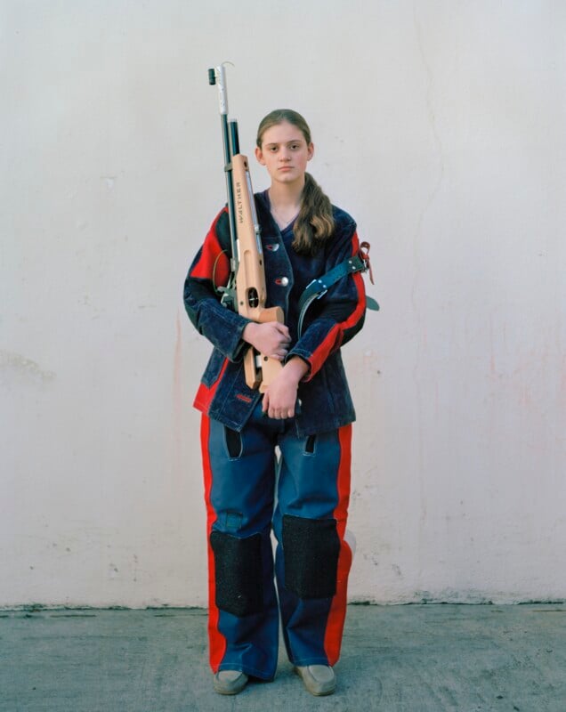 A young person in a blue and red shooting suit stands against a plain wall, holding a Walther air rifle vertically.