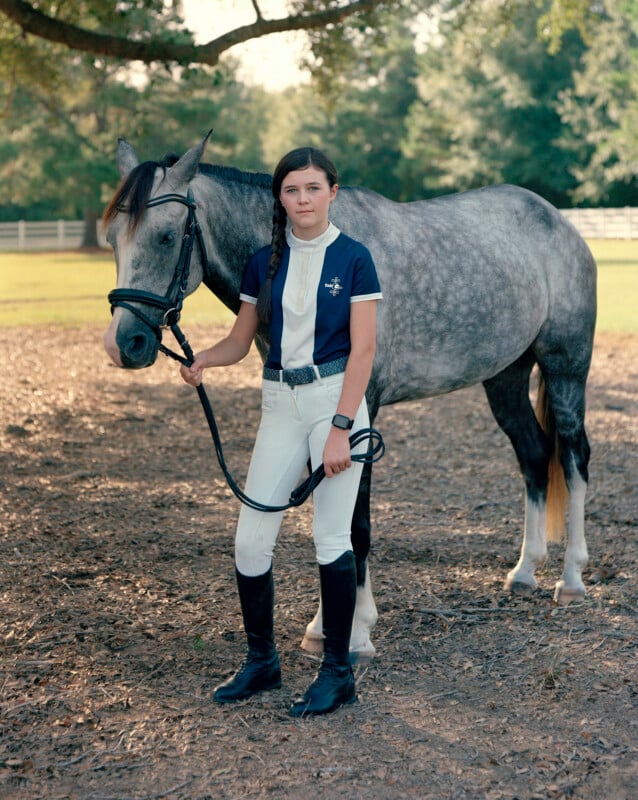 A young girl with a side braid stands next to a grey horse, holding its reins while wearing equestrian riding gear.